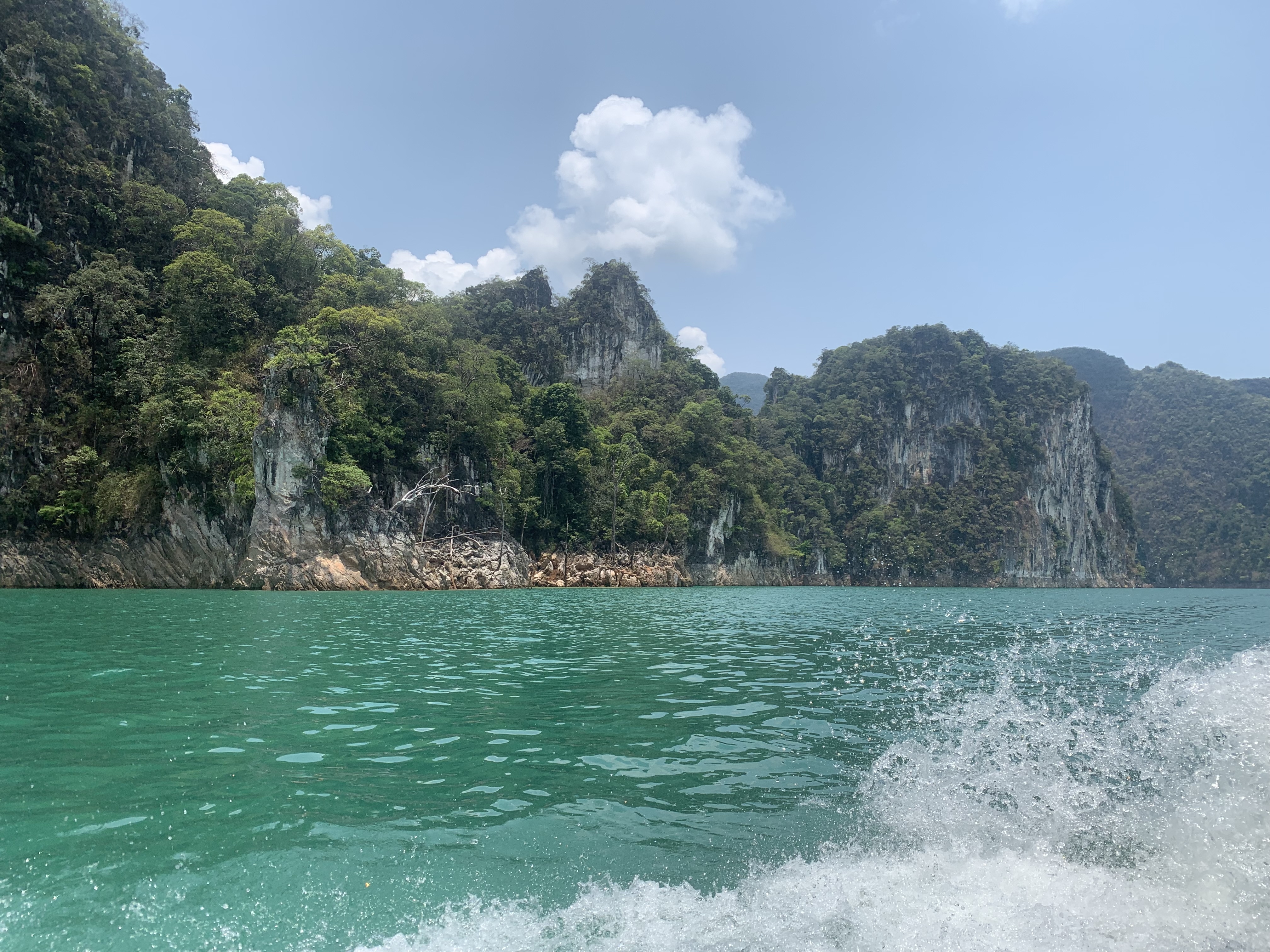 Zicht vanaf een varende boot over turquoise water met opspattend schuim richting beboste kalkstenen karstbergen.